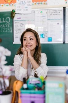 A teacher seated at their desk smiling, in front of a blackboard full of notes