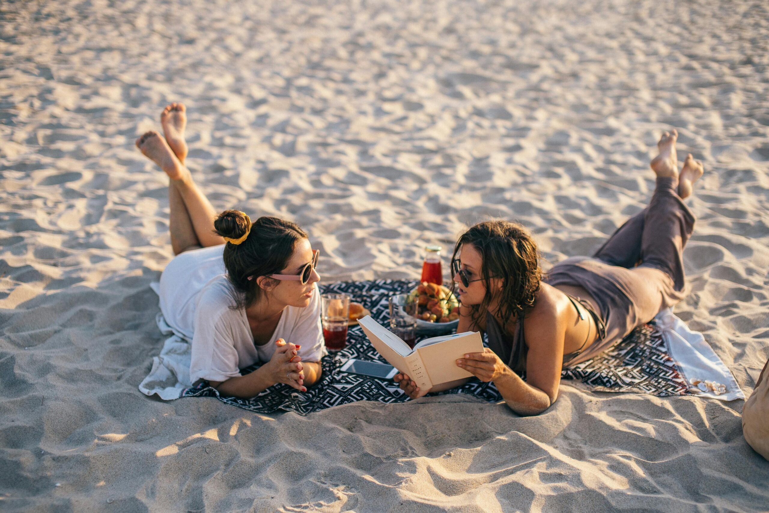 Two people lie on a blanket on a sandy beach, enjoying a relaxing beach day. One holds a book open in their hand, and they chat, perhaps about the book