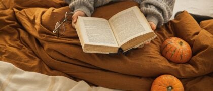 A person in bed reading a book amid cozy blankets. Beside them are a pair of glasses and a few decorative gourds.