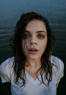 A woman with wet hair and clothing, smeared eye makeup, and a dazed expression stands in front of a body of water