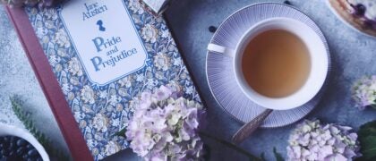 A tableau featuring Jane Austen's "Pride and Prejudice," fresh purple hydrangeas and greenery, a cup of tea, a blueberry cake, and scattered blueberries, creating an inviting scene for reading