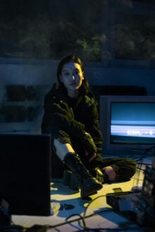 A woman wearing combat boots, seated on the floor of a semi-dark room. She is surrounded by computer monitors and cables.