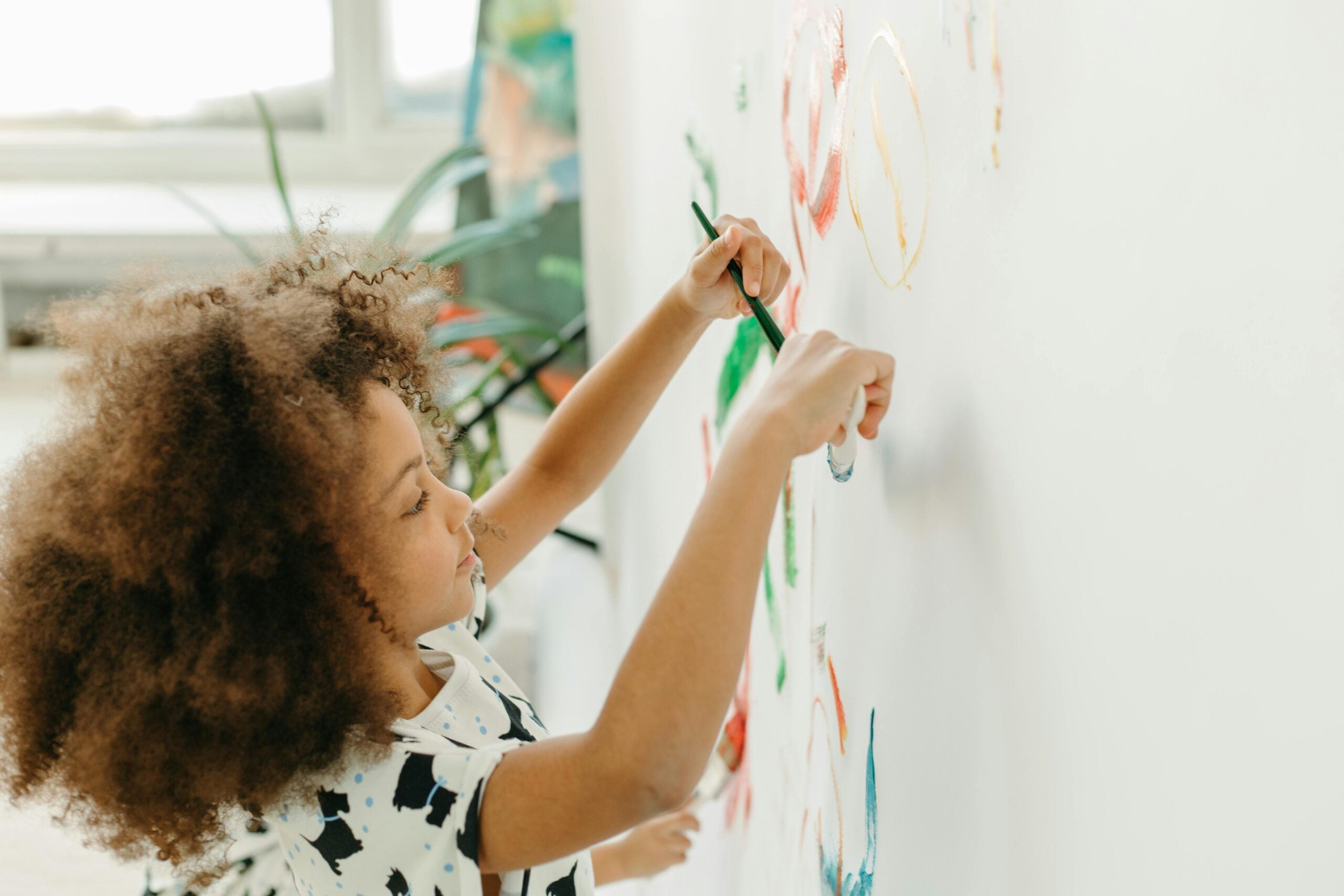 A young girl with curly hair joyfully painting colorful designs on a wall, showcasing her creativity and artistic expression.