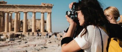 Person with long hair and yellow backpack takes a photo of ancient Greek ruins under a clear blue sky.