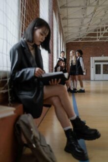 A young person sits alone on a bench writing in a notebook. A group of other young people is standing in the background by the wall of a school gym, looking at the person in the foreground.